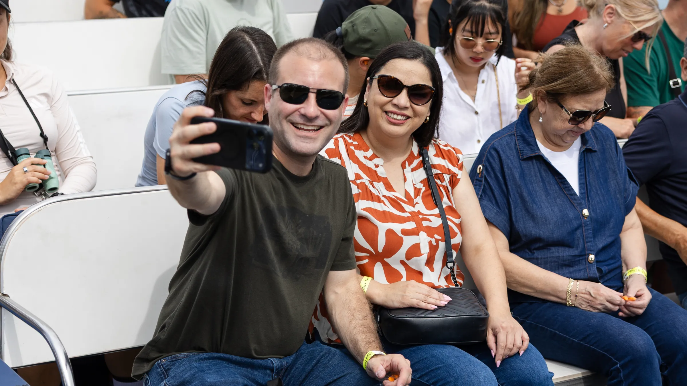 Man and woman taking a selfie, sitting on a bench in a crowd.