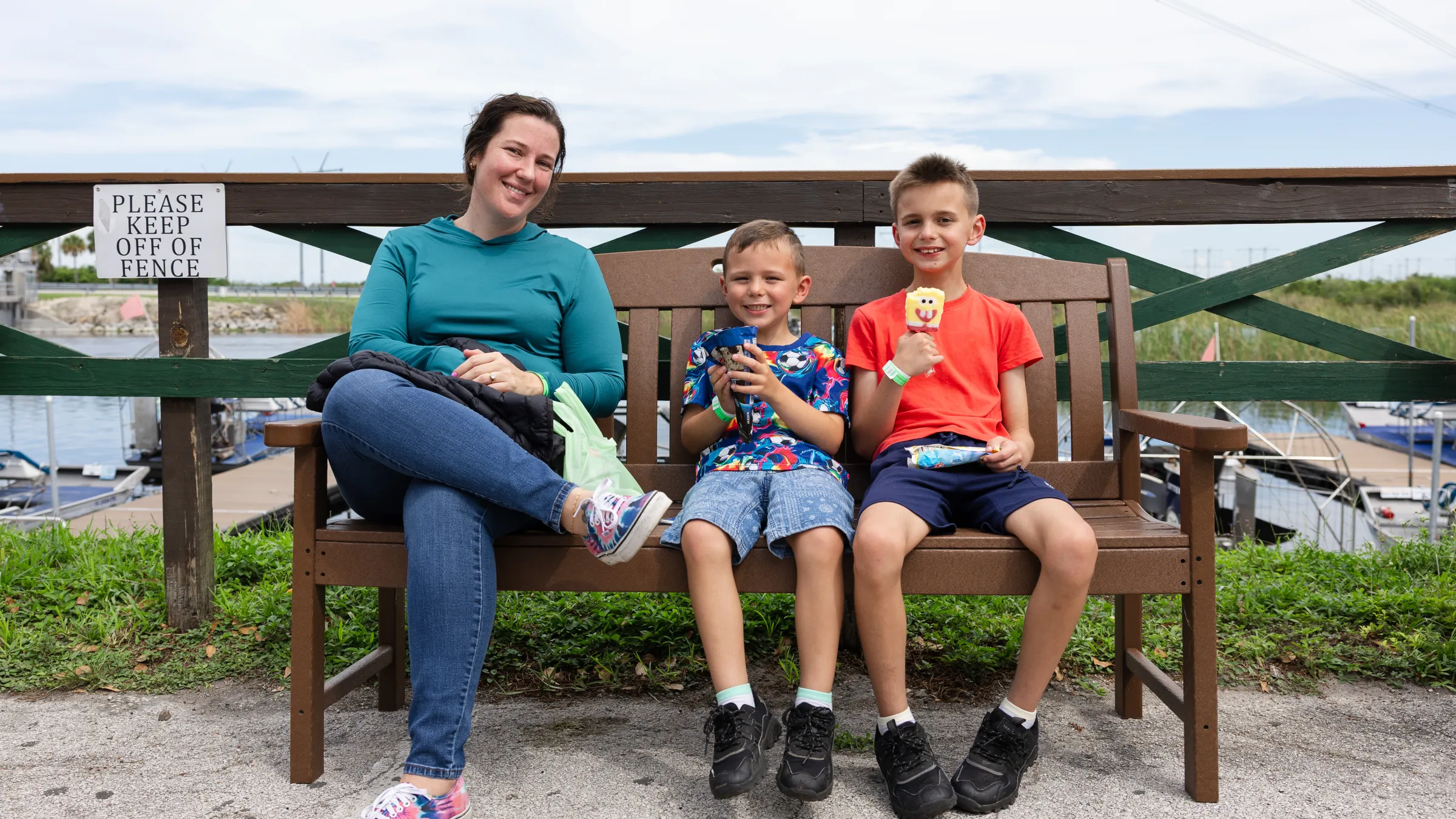 Person and two kids sitting on a bench, holding ice creams near a fenced area.