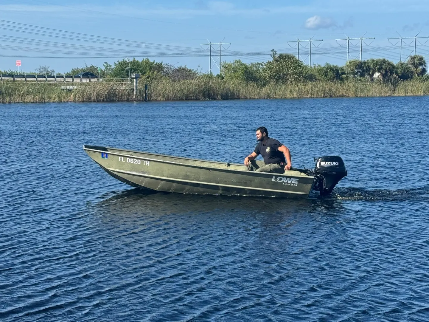Person in a small metal boat on a large body of water, near reeds and power lines under clear blue sky.