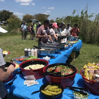 People serving food at an outdoor buffet with salads and snacks on a sunny day.