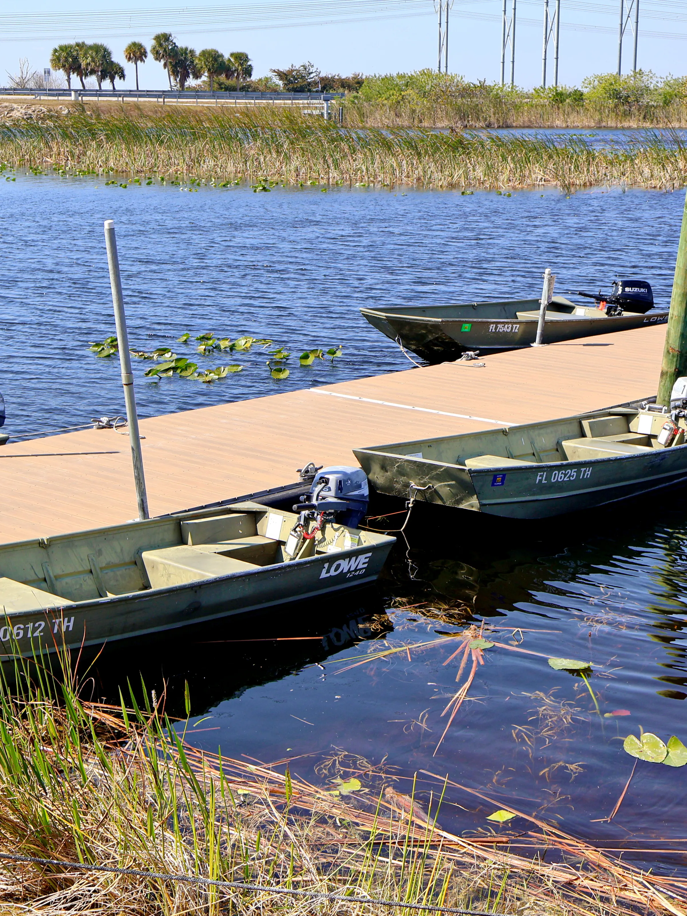 Small motorboats docked at a pier on a calm lake with reeds in the background.