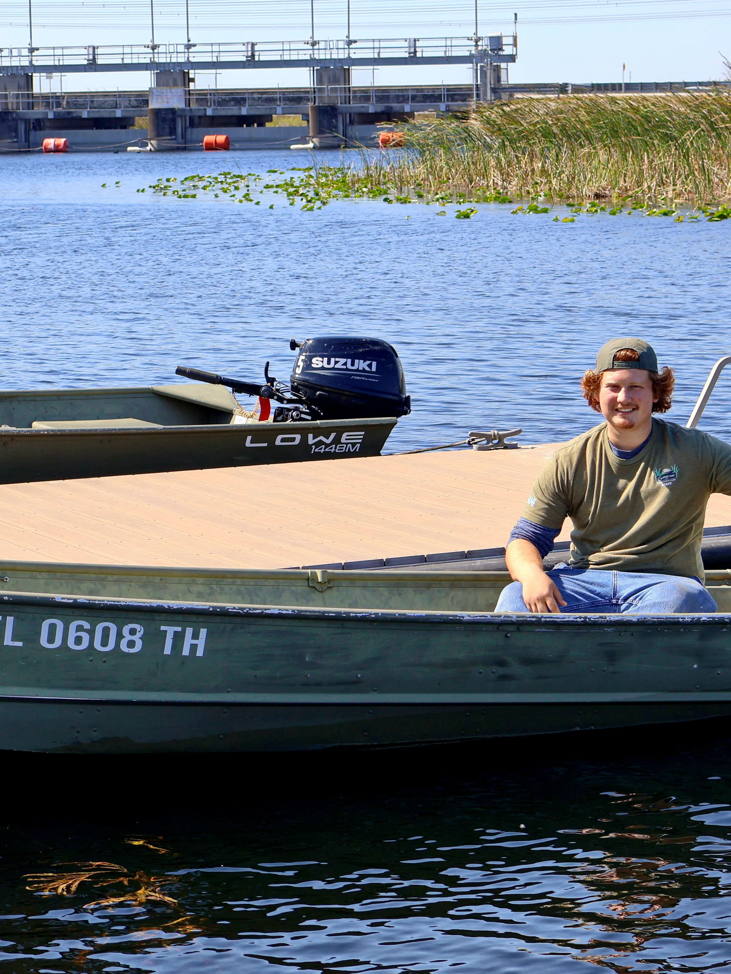 Person sitting on a small green boat docked on a lake with reeds and a structure in the background.