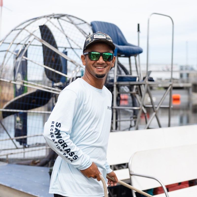Man in sunglasses smiles by an airboat on a sunny day.